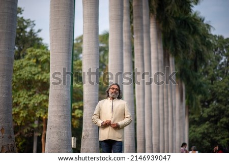 A grey bearded man in fashionable jacket posing for picture  while standing amid trees in line. 