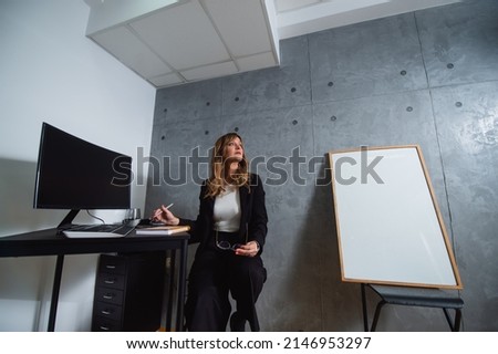 A wistful senior caucasian woman sits on a chair and looks out the window while her right hand is on the desk and in her left hand are glasses.The whiteboard for presentation leans against a gray wall
