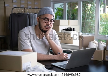 Online seller entrepreneur in a warehouse looking at laptop and worrying about e-commerce business due to lack of sales and customers.