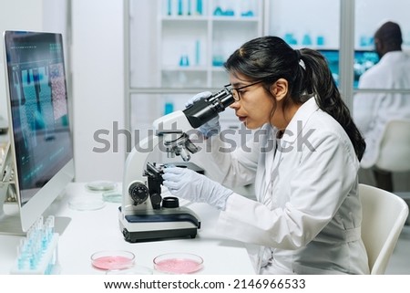 Side view of female biochemist in whitecoat, gloves and eyeglasses looking in microscope while studying virus in laboratory Royalty-Free Stock Photo #2146966533