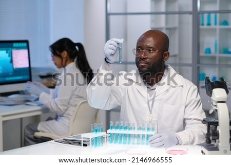 African American male virologist in gloves and lab coat looking at blue liquid in flask against female colleague working in front of computer Royalty-Free Stock Photo #2146966555