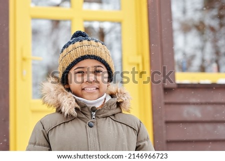 Contemporary cute intercultural boy in winterwear standing in front of camera against new country house of his family in the forest