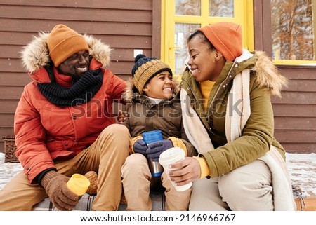 Cute happy boy in winterwear sitting between his parents on porch of their family country house, having hot tea and chatting