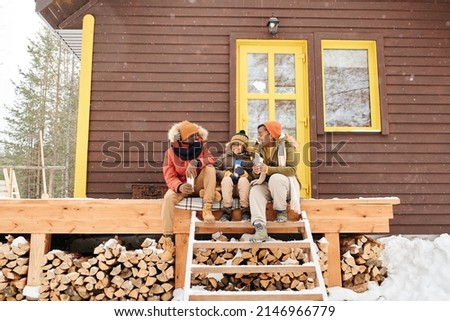 Happy young family of three sitting on wooden porch by their countryhouse, having hot tea and talking on winter day Royalty-Free Stock Photo #2146966779