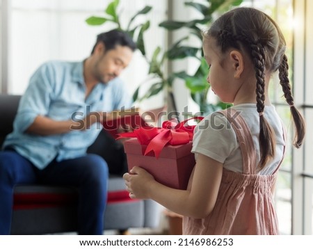 Selective focus, cute daughter holding red box gift with blurry background of father reading book on sofa. Girl want to surprise dad on special day. Father's day or Birthday concept.