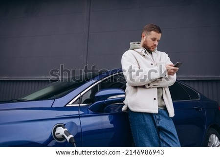 The guy sat down on the hood of the car. His car is charging at the charging station. A man looks at the smartphone screen and smiles.