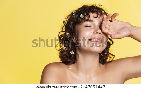 Natural beauty and skincare. Portrait of happy woman with curly hair and healthy face without acne or makeup, chamomile in hair, touching skin and smiling at camera, yellow background Royalty-Free Stock Photo #2147051447