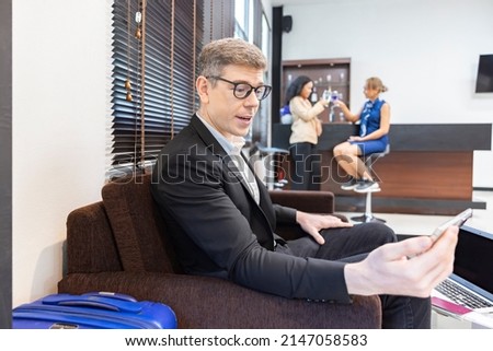 Business executive making a video call by mobile phone,  having fun with device while waiting for his flight at airport lounge and blurred background passengers at bar counter. Focus on his face  
