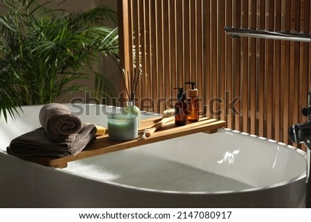 Wooden bath tray with candle, air freshener and bathroom amenities on tub indoors Royalty-Free Stock Photo #2147080917