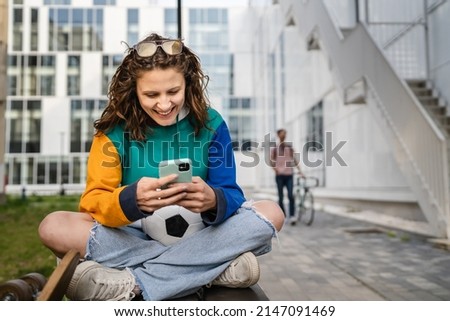 One young caucasian woman female sitting on the bench in front of the building or at stadium with soccer ball waiting for the football game using mobile phone real people copy space Royalty-Free Stock Photo #2147091469
