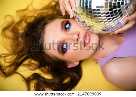 A beautiful woman with bright makeup on a yellow background in sports purple clothes holds a disco ball in her hands. photo in the style of the 80s