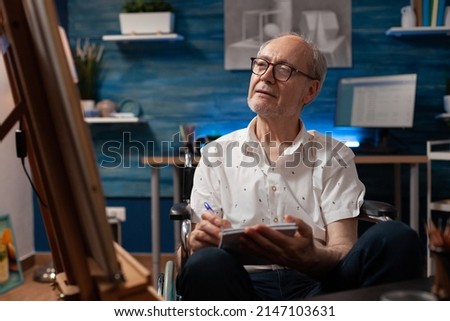 Elderly man artist living with disability doing sketch on notebook for practice before drawing on easel in home art studio. Creative senior hobby art creator taking notes keeping journal.