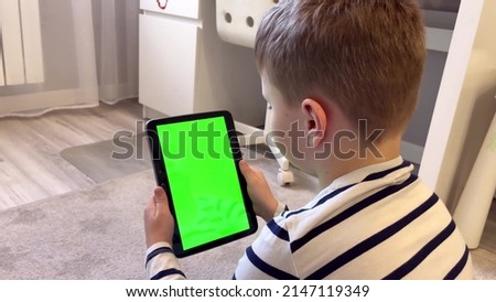 Child is sitting on the carpet and using a digital tablet PC with green screen, back view. Boy holding a tablet with green screen in hand