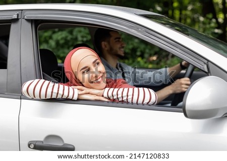 Smiling millennial arab female in hijab ride in car looks out the open window outdoor, enjoy journey, man drives car. Travel in summer, weekends on road, holidays with family after covid-19 pandemic Royalty-Free Stock Photo #2147120833