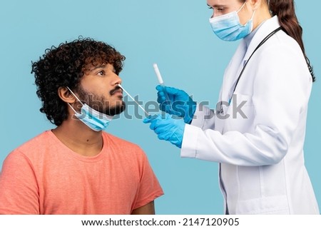 Female doctor in protective face mask taking nasal speciment from young indian man, using cotton swab, wearing medical gloves, blue studio background, closeup. COVID-19 diagnostic concept Royalty-Free Stock Photo #2147120905