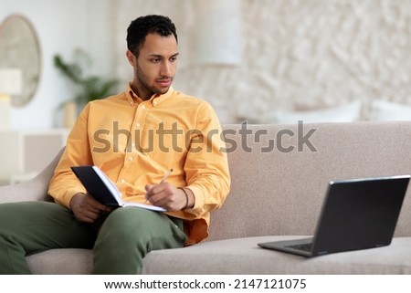 Portrait of young focused Arabic man using laptop sitting on couch, writing in notebook. Serious millennial guy browsing internet, watching webinar studying online, taking notes, free copy space