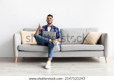 I Like. Portrait Of Happy Excited Middle Eastern Man Sitting On Couch Using Laptop And Showing Thumbs Up Sign Gesture, Working Or Studying Remotely, Posing To Camera In Living Room, Full Body Length Royalty-Free Stock Photo #2147121243