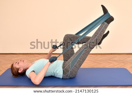 Middle-aged woman doing pilates exercises with rubber resistance band on the mat at home. Fitness, sport, training and healthy lifestyle concept.