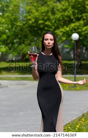 Attractive lady in black elegant dress touching neck and enjoying red wine on sunny summer day in park