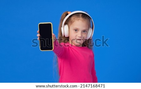 A cute girl in a bright pink sweater holds a mobile phone in her hands and listens to music in wireless headphones. The concept of children's applications on gadgets