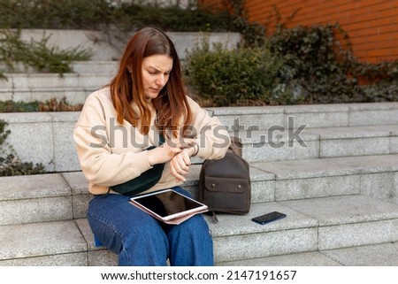 Woman looking at clock an waiting something. Cheerful young femile sitting on stairs on city street, Urban lifestyle concept. Traveler