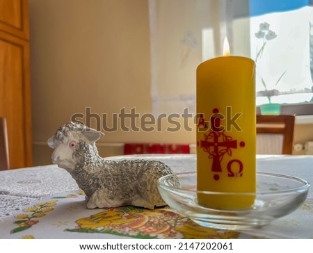 A modest lamb and a lit candle with alpha and omega signs standing on the table as Easter symbols.