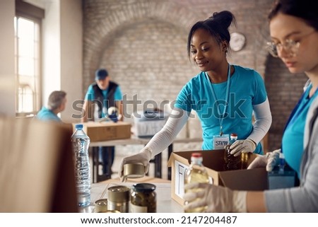 Young happy African American woman preparing donation boxes while volunteering with group of people  at food bank. Royalty-Free Stock Photo #2147204487