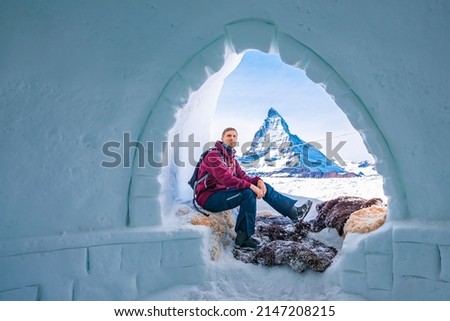 Portrait of young tourist resting at entrance of igloo. Famous snowcapped matterhorn peak is in background. Man enjoying winter in alpine region.