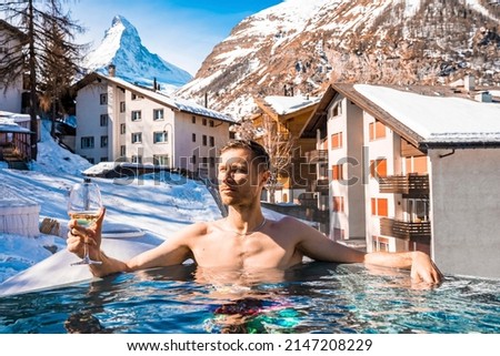 Contemplating man enjoying wine while swimming in pool. Tourist relaxing against snow covered matterhorn mountain. Scenic view of townscape during winter.