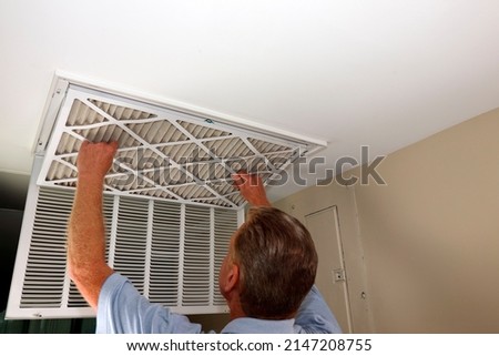 Dirty air filter being removed from a home furnace air intake vent by an adult white male close-up. Adult white male removing a dirty air filter from a furnace air duct intake vent in a home ceiling. Royalty-Free Stock Photo #2147208755