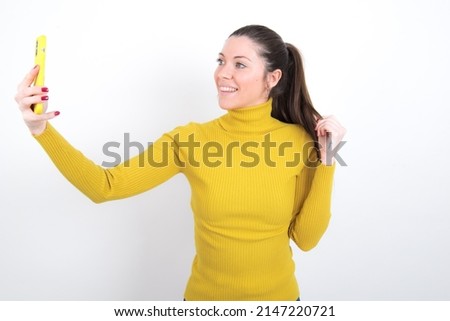 young caucasian girl in yellow turtleneck over white background  smiling and taking a selfie ready to post it on her social media.