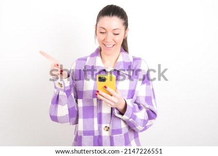 Smiling young caucasian woman wearing plaid shirt over white background pointing finger at blank space holding phone in one hand