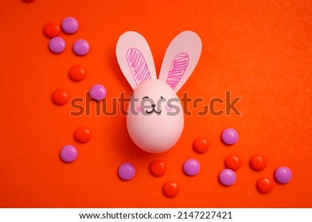 Greeting card for Easter. Easter egg with bunny ears on a red background with small colorful candies. Rabbit face on a chicken egg.