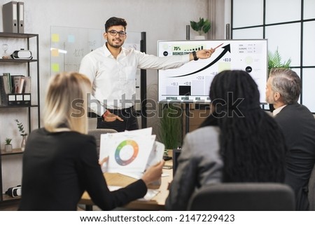 Middle aged male bearded indian chief showing company's work infographics on digital wall screen during meeting with team of diverse multiracial male and female businesspeople. Royalty-Free Stock Photo #2147229423