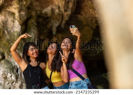 Young Asian woman friends using mobile phone taking selfie together while travel in the cave on island beach lagoon in sunny day. Happy female enjoy and fun outdoor lifestyle on summer vacation trip