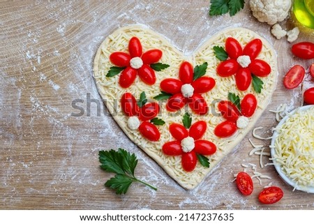 Heart shaped pizza dough covered with mozzarella cheeses,flower shaped tomatoes,cauliflowers and parsley on wooden cutting board background.Love vegetarian concept for Valentine Day or Mother Day
