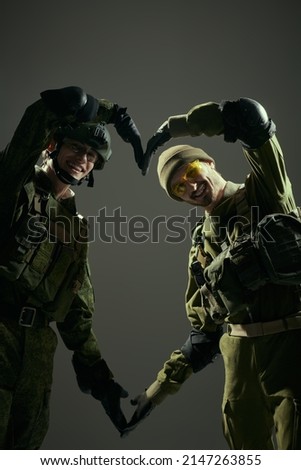 Two Army soldiers in Protective Combat Uniform hold their arms in the shape of heart and smile. Khaki background. Comrades in arms, friendship. Military concept.