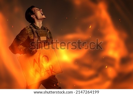 No War concept. Angry soldier in Protective Combat Uniform holds a sign "No War" screaming furiously in a dark background engulfed in flames. Studio shot. Royalty-Free Stock Photo #2147264199