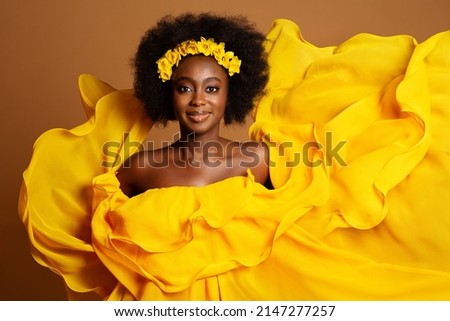Happy Dark Skinned Woman in Yellow Fashion Dress. Beauty Afro American Model dancing in Silk Gown waving Flying on Wind over Beige Background. Yellow Flower Wreath in Women Black Curly Hairstyle Royalty-Free Stock Photo #2147277257