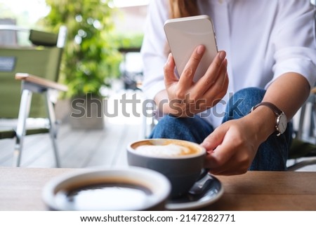 Closeup image of a young woman holding and using mobile phone while drinking coffee with friend in cafe