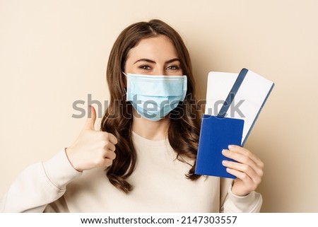 Travel and covid-19 pandemic. Happy woman tourist in medical mask, showing thumbs up and passport with two tickets, beige background