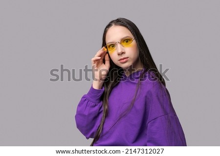 Portrait cute teenage girl in purple sweatshirt and yellow glasses posing in the studio on a gray background. Caucasian girl raotae on camera