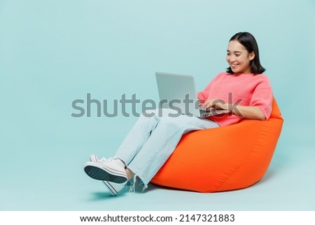Full body young smiling happy woman of Asian ethnicity 20s wear pink sweater sit in bag chair hold use work on laptop pc computer isolated on pastel plain light blue color background studio portrait. Royalty-Free Stock Photo #2147321883