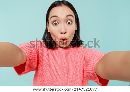 Close up surprised young amazed woman of Asian ethnicity 20s in pink sweater doing selfie shot pov on mobile phone isolated on pastel plain light blue color background studio. People lifestyle concept