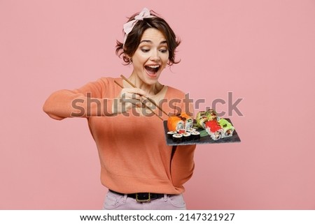 Young excited amazed cool fun woman 20s in casual clothes hold in hand eat makizushi sushi roll served on black plate traditional japanese food chopsticks isolated on plain pastel pink background. Royalty-Free Stock Photo #2147321927