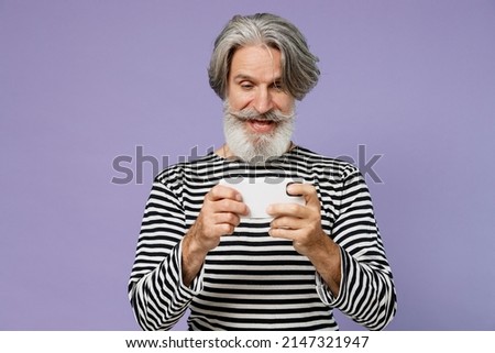 Elderly gambling gray-haired mustache man 50s in striped turtleneck using play racing app mobile cell phone hold gadget smartphone for pc video games isolated on plain pastel light purple background