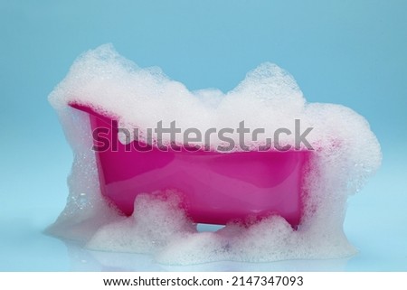 Toy bathtub overflowing with foam on light blue background Royalty-Free Stock Photo #2147347093