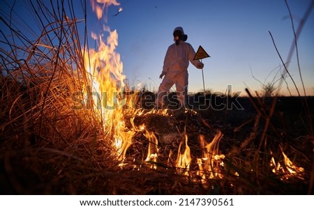 Fireman ecologist fighting fire in field at night. Man in protective radiation suit and gas mask near burning grass with smoke. Natural disaster concept.