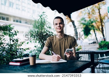 Content male browsing smartphone and looking at camera while taking notes in notepad at table with coffee on terrace of cafeteria on blurred background