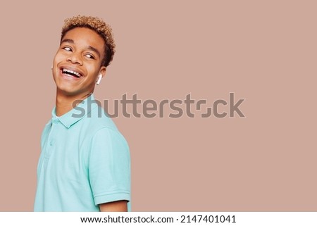 Glad young afro latin american man feels great, wears in green shirt, listens to a new exciting podcast, broadly smiles and looks away stands over beige background.